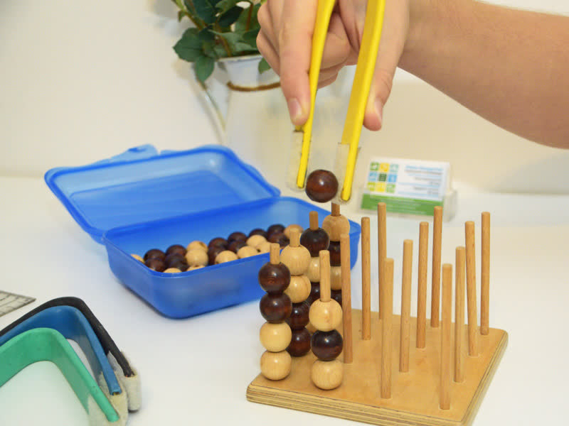 Greifzange hält eine Holzkugel über einem Steckspiel mit Holzstäben, daneben eine geöffnete Box mit weiteren Kugeln